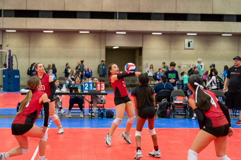 A volleyball game is in progress with a player in red preparing to hit the ball. The scoreboard reads 21-13. Other players are on the court and spectators are visible in the background.