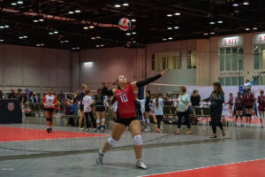 A volleyball player in a red jersey prepares to serve during an indoor game with spectators and other players visible in the background.