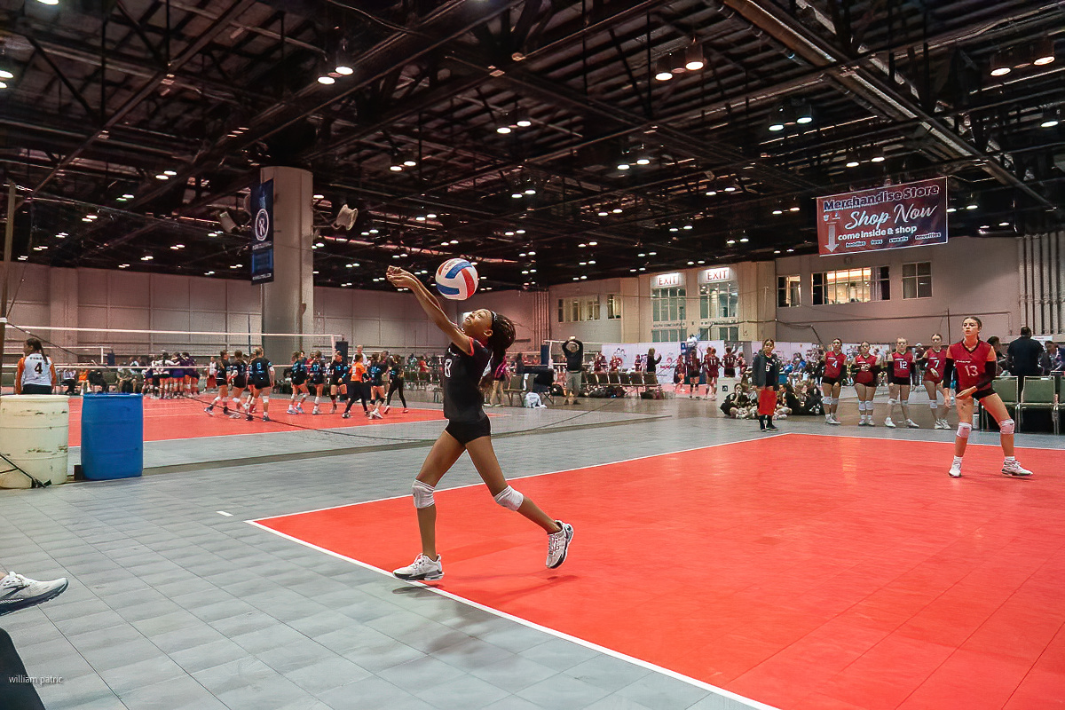 A volleyball player prepares to serve in an indoor gymnasium, with other players and teams visible in the background.