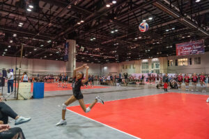 A volleyball player in a black uniform prepares to bump the ball in an indoor court during a match, with spectators and teams visible in the background.