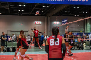 A volleyball player in a red jersey jumps for a spike as teammates and opponents watch. A score of 4 to 1 is displayed, and spectators and coaches observe from the sidelines.