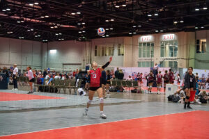 A volleyball player in a red jersey prepares to serve the ball on an indoor court during a match. Other players and spectators are visible in the background.