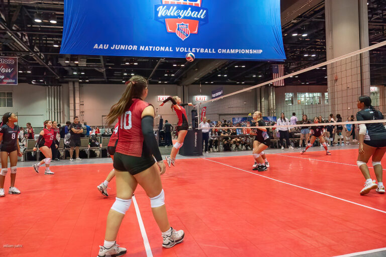 A volleyball game in progress at the AAU Junior National Volleyball Championship with players in red and green uniforms actively engaged on a court with a red floor.