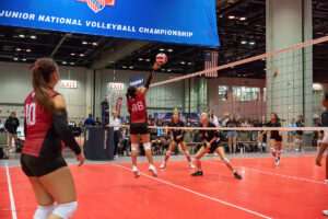 A volleyball player jumps to spike the ball during a match in a large indoor stadium. Other players, wearing red and black uniforms, prepare to receive the ball. An event banner is seen in the background.