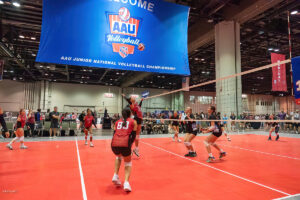 A youth volleyball team in red jerseys faces off against a team in black jerseys during a match at the AAU Junior National Volleyball Championship in an indoor court.