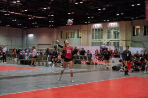 A volleyball player wearing a red jersey with the number 23 serves the ball in an indoor volleyball court during a match. Other players and spectators are visible in the background.