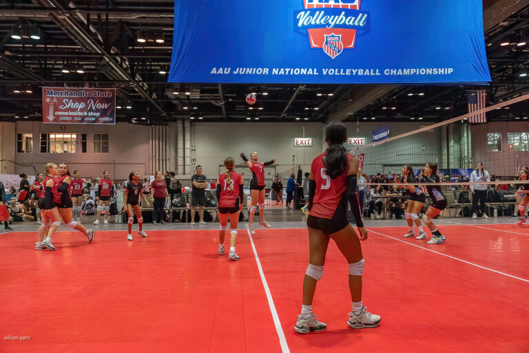 Indoor volleyball game in progress with teams competing at the AAU Junior National Volleyball Championship. Players in red and black uniforms are on the court, with a large scoreboard overhead.