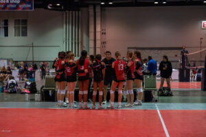 A volleyball team in red uniforms huddles around their coach on an indoor court, preparing for a game.