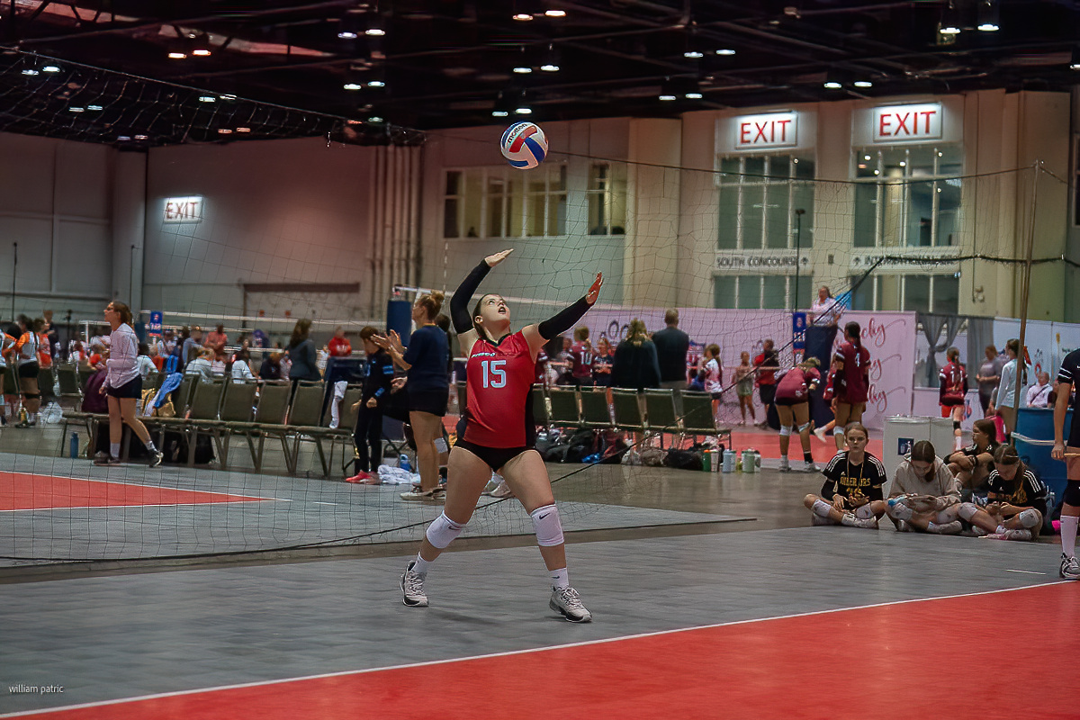 Indoor volleyball player wearing a red jersey with the number 15 prepares to serve the ball during a match. Spectators and teammates are visible in the background.