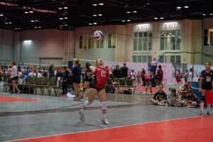 A volleyball player wearing a red jersey with the number 15 prepares to serve the ball during an indoor match, with teammates and spectators in the background.