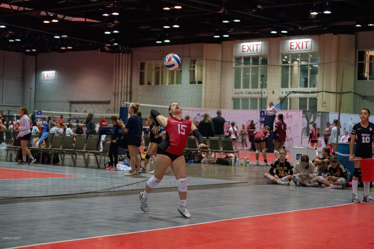 A volleyball player wearing a red jersey with the number 15 prepares to serve the ball during an indoor match, with teammates and spectators in the background.