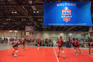 Indoor volleyball game in progress with participants in red jerseys under a large "AAU Volleyball" banner at the AAU Junior National Volleyball Championship.