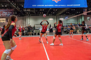 A volleyball player prepares to set the ball during a match at the AAU Junior National Volleyball Championship. Other players and a net are visible on the court.