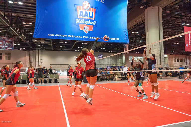 A volleyball player in a red jersey jumps to spike the ball over the net during a match. Four players in blue jerseys are on the opposing side. A large blue banner for the AAU Junior National Championship is hanging.