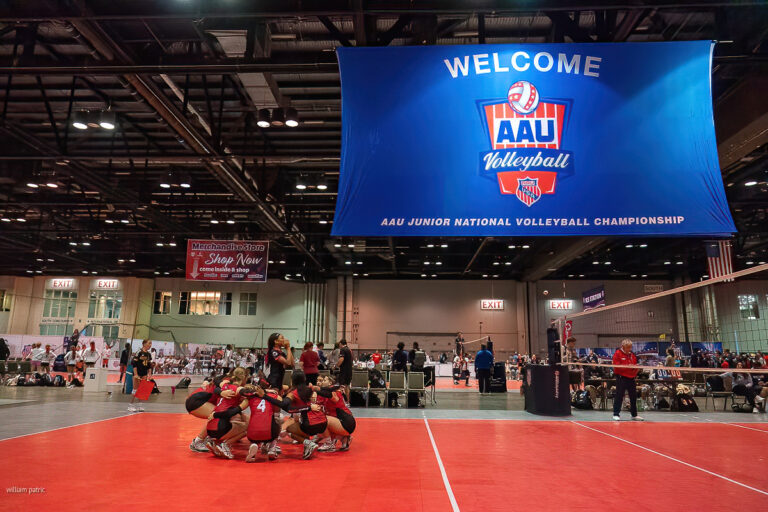 A volleyball team huddles on a red court beneath a large sign that reads, "Welcome AAu Volleyball, AAU Junior National Volleyball Championship." Spectators are visible in the background.