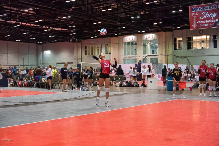 A volleyball player in a red jersey is serving the ball during an indoor match. Other players and spectators are visible in the background, along with a scoreboard and advertising banners.