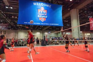 Indoor volleyball game at the AAU Junior National Volleyball Championship; players in action on the court, with a large blue event banner hanging overhead.