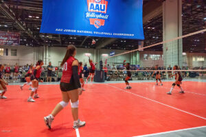 A women's volleyball match is in progress at the AAU Junior National Volleyball Championship. Players are actively engaging near the net with a large tournament banner overhead.