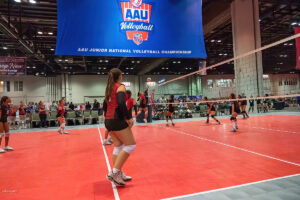 A volleyball game in progress at the AAU Junior National Volleyball Championship. Players in red and black jerseys are on the court, with a large AAU Volleyball banner overhead.