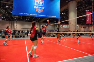 Indoor junior volleyball match in progress, with players on a red court under a large AAU Junior National Volleyball Championship banner. Player in red uniform jumps to block or spike the ball.