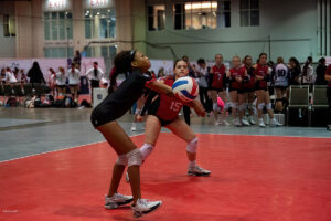 Two volleyball players, one in a black jersey and the other in a red jersey, prepare to receive the ball in an indoor court. Other players and the audience are visible in the background.