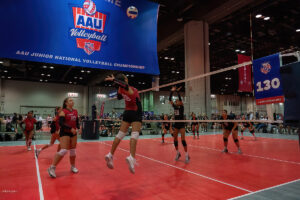 A youth volleyball game in progress at the AAU Junior National Volleyball Championship, featuring players in red and black jerseys performing various actions on the court.