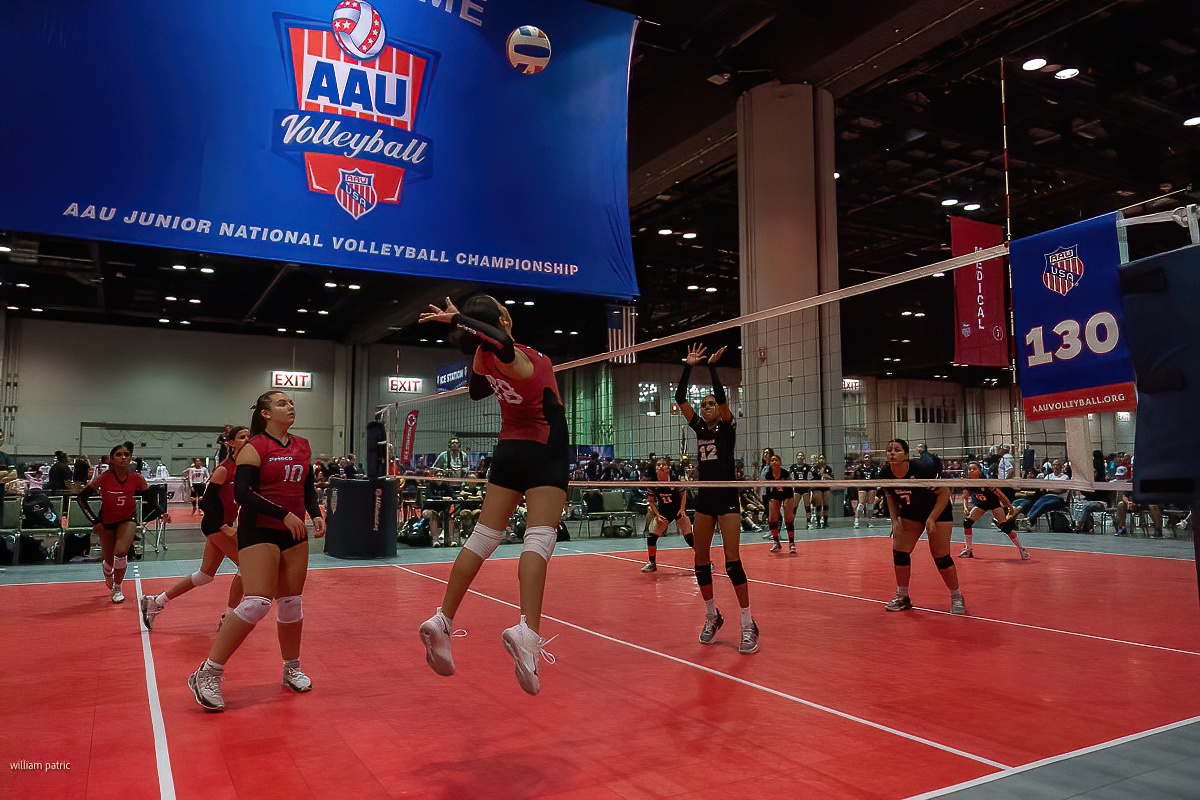 A youth volleyball game in progress at the AAU Junior National Volleyball Championship, featuring players in red and black jerseys performing various actions on the court.
