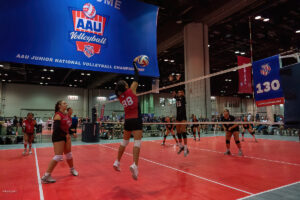 Indoor volleyball game with players mid-action; one player in a red jersey jumps to spike the ball, while opposing players in black jerseys prepare to block. A large AAU Volleyball banner hangs overhead.