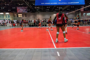 Indoor volleyball match with female players on a red court during the AAU Junior National Volleyball Championship. Some players are preparing for a play while others are in action.