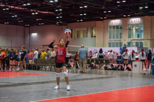 A volleyball player in a red jersey prepares to serve the ball during an indoor match, with spectators and other players in the background.