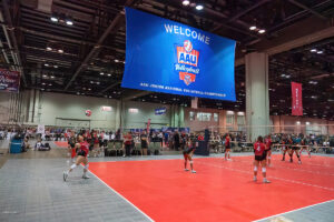 Indoor volleyball game with two teams competing at the AAU Junior National Volleyball Championship, featuring a large blue banner reading "Welcome" and seats filled with spectators in the background.