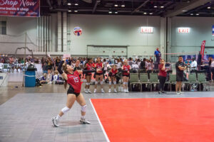 A volleyball player wearing number 15 serves the ball during an indoor match while her teammates look on from the sideline.