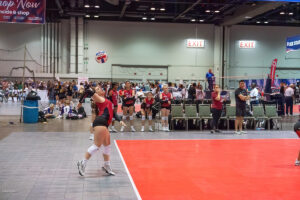Indoor volleyball game in progress; one player prepares to hit the ball while teammates and coaches watch from the sideline.