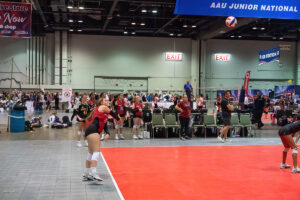 Indoor volleyball players preparing for a serve on a red court during a tournament with spectators and other players in the background. Banner reads "AAU Junior National".