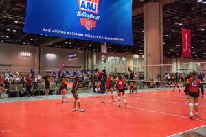 Indoor volleyball match in progress at the AAU Junior National Volleyball Championship. Players in red and black uniforms are on the court with spectators and banners in the background.