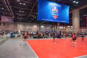 Indoor volleyball tournament in progress with players in red uniforms on a red court, and a large banner overhead reading "Welcome AAU Volleyball" at the AAU Junior National Volleyball Championship.