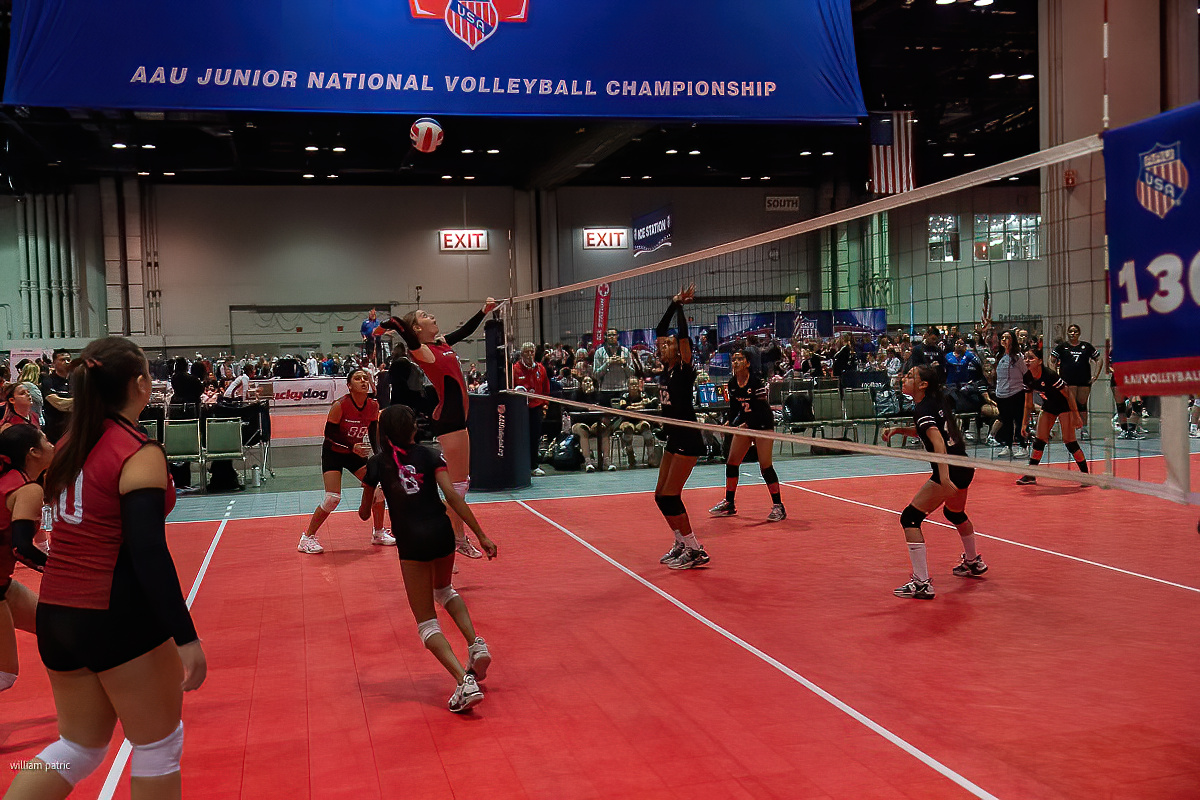 Indoor volleyball game in progress during the AAU Junior National Volleyball Championship with players preparing to receive a serve. The court is red with a blue banner overhead.