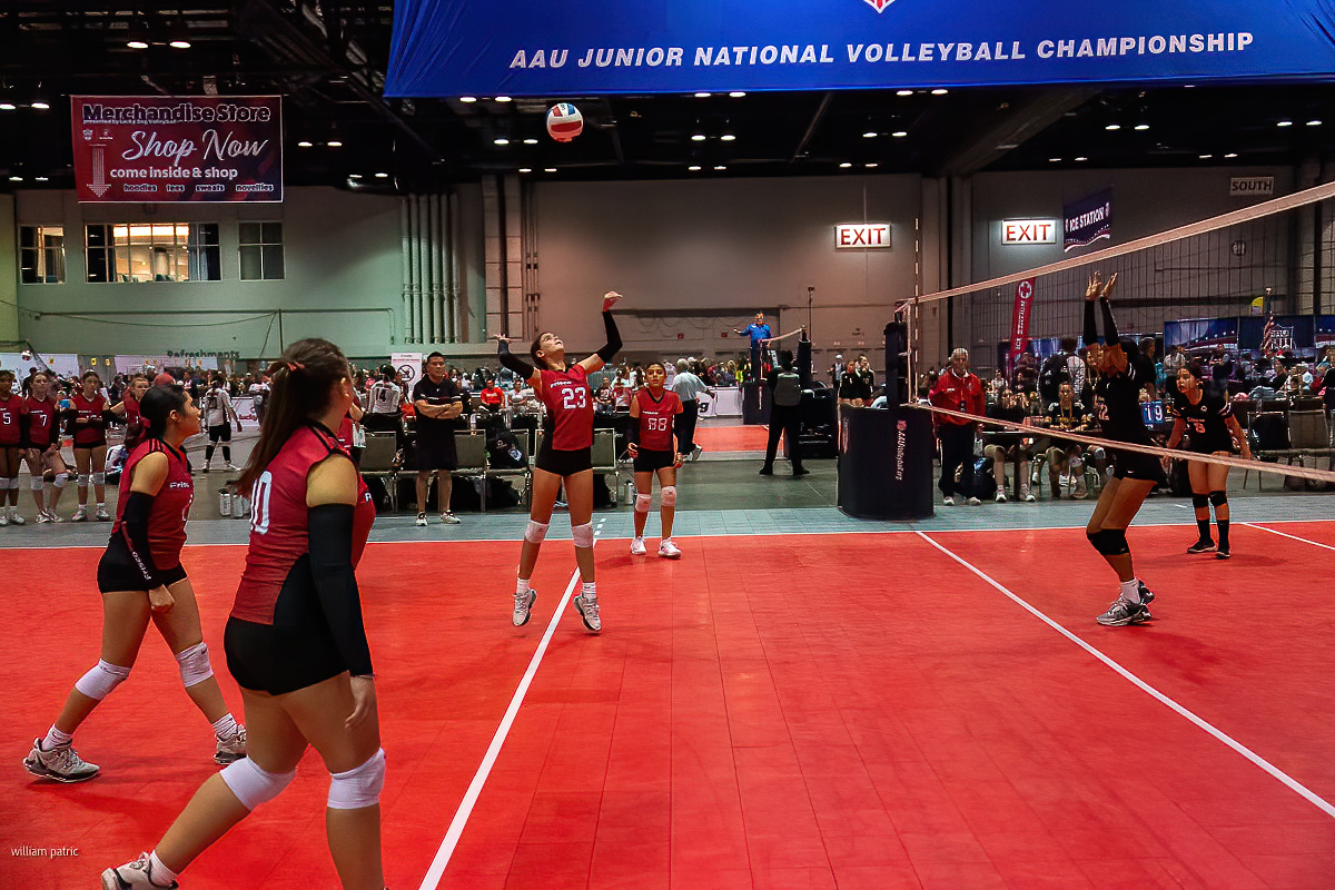 A girls' volleyball team is playing an indoor match at the AAU Junior National Volleyball Championship, with one player preparing to hit the ball over the net.