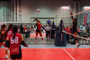 A volleyball player wearing a red jersey jumps to hit the ball while an opponent attempts to block at the net. Teammates and spectators are in the background.