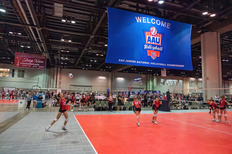 Indoor volleyball game in progress at the AAU Junior National Volleyball Championship event. Players in red uniforms are on the court with spectators in the background. Large welcome banner overhead.