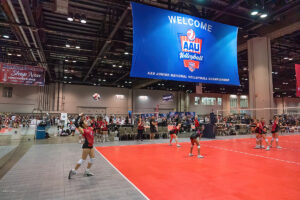 A volleyball game taking place indoors, with players in red and black uniforms. A large banner above reads "Welcome AAU Junior National Volleyball Championship." Spectators are seated in the background.