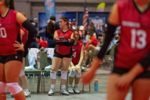 Several volleyball players wearing red jerseys stand and sit in a sports hall. One player, number 4, stands near a seated teammate. The background features banners, flags, and spectators.