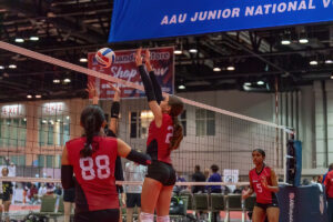 A volleyball player jumps to block the ball at the net during a match, while another player looks on. The event banner above reads "AAU Junior National.