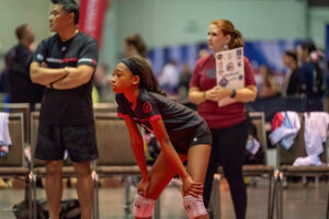 A young volleyball player leans forward with hands on knees, appearing focused. Two individuals, one with crossed arms and another holding a booklet, stand in the background. Several chairs are nearby.