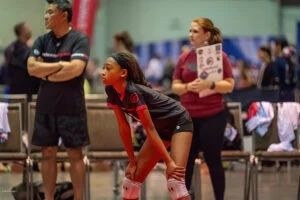 A young volleyball player leans forward with hands on knees, appearing focused. Two individuals, one with crossed arms and another holding a booklet, stand in the background. Several chairs are nearby.