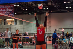 A volleyball player in a red jersey with the number 10 jumps to set the ball, while another player in a black jersey watches. The scene takes place in an indoor sports facility.