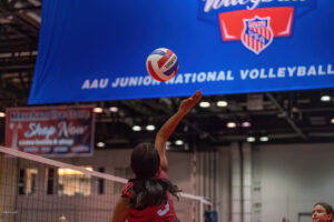A volleyball player in a red uniform jumps to spike the ball during the AAU Junior National Volleyball tournament, as a large blue banner hangs in the background.