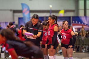 A volleyball coach discusses strategy with his team of players in red jerseys during a game. Players look attentive and focused.