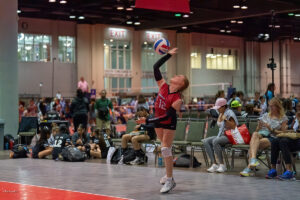 A volleyball player in a red jersey serves the ball in an indoor sports arena, with spectators and other players visible in the background.