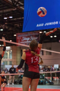 A volleyball player wearing a red jersey with the number 13 jumps to hit the ball over the net during a game at an indoor court.
